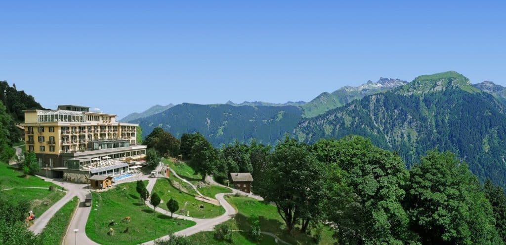Märchenhaftes Berghotel auf grünem Hügel mit Panoramablick auf die Alpen unter strahlend blauem Himmel.