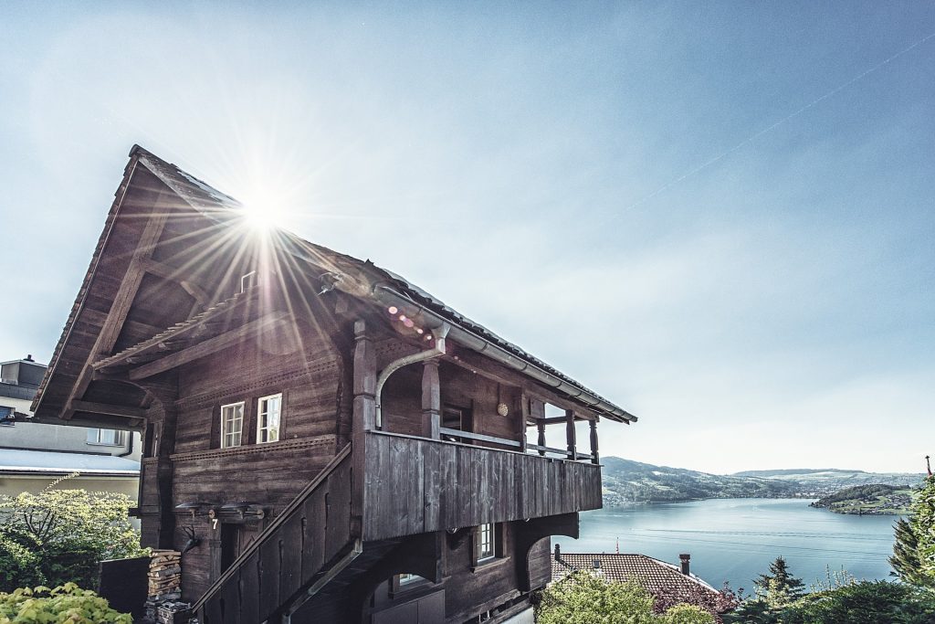 Traditioneller Spycher aus Holz mit Blick auf einen See, umgeben von Hügeln und Bäumen bei strahlendem Sonnenschein.