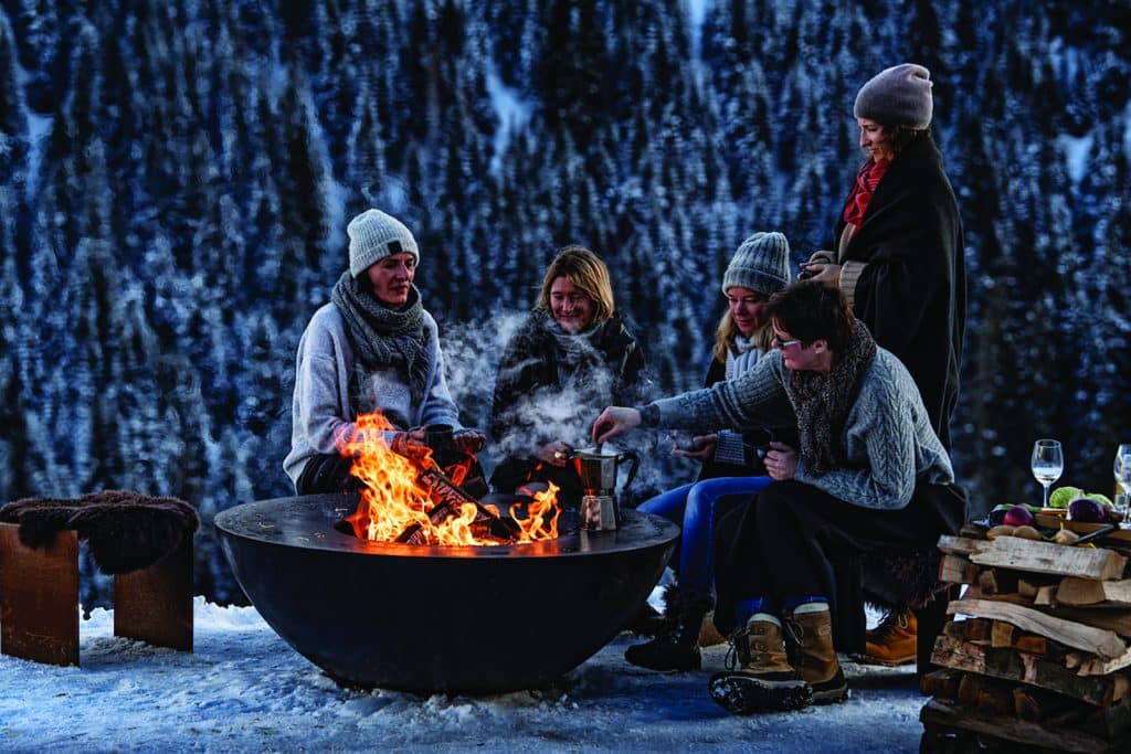 Gruppe von Menschen am Feuerplatz im Schnee – winterliches Outdoor-Erlebnis mit Stil und Gemütlichkeit in der Hotellerie.