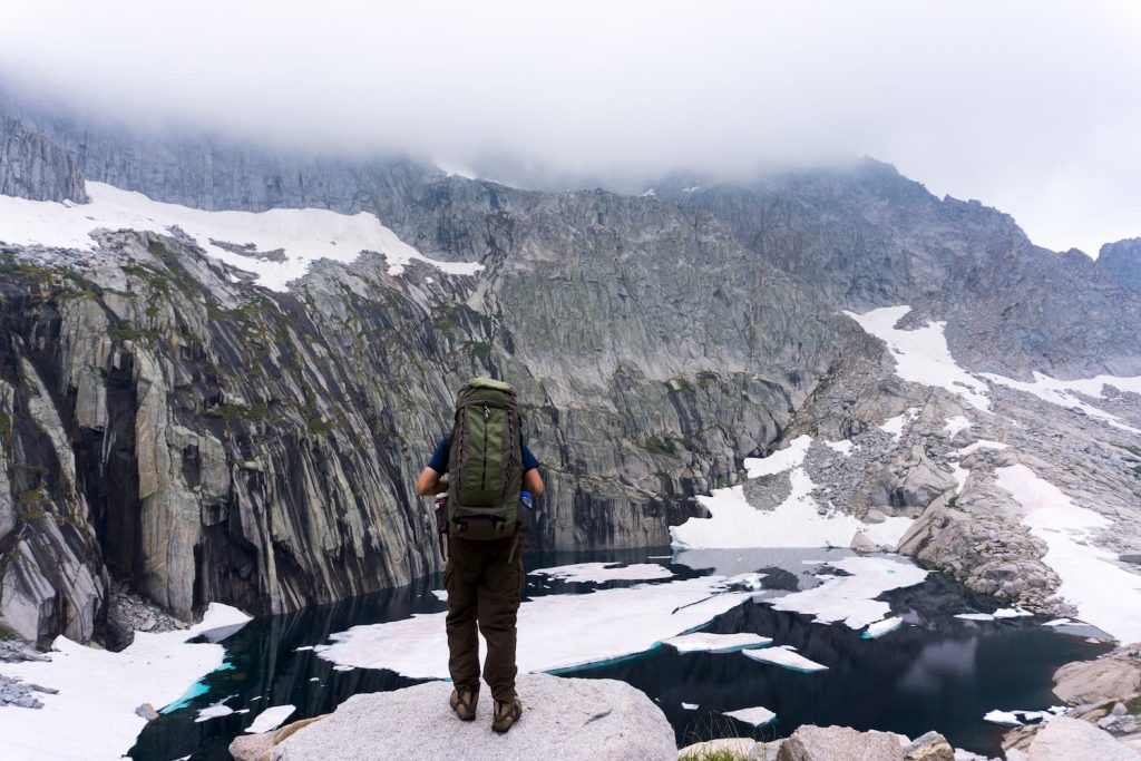 Wanderer mit Rucksack auf Felsen vor verschneiter Berglandschaft – Symbolbild für Winterurlaub im neuen Normal.