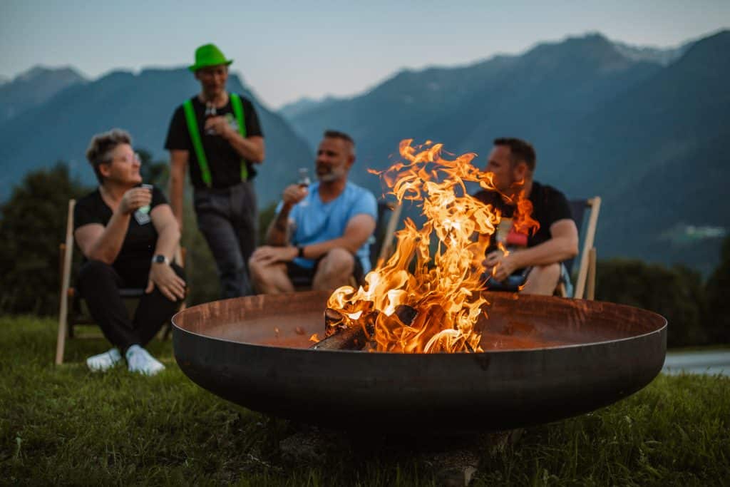 Abendliche Stimmung mit einer brennenden Feuerschale. Eine Frau und 3 Männer sitzen lachend um die Feuerschale und stoßen mit einem Bier auf diesen großartigen Moment an.