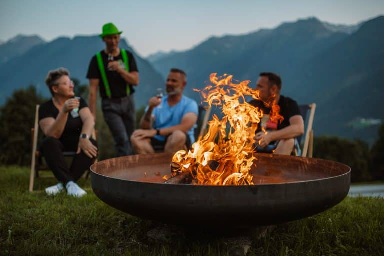 Abendliche Stimmung mit einer brennenden Feuerschale. Eine Frau und 3 Männer sitzen lachend um die Feuerschale und stoßen mit einem Bier auf diesen großartigen Moment an.